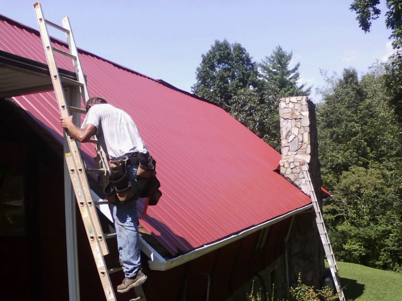 Roofer on a ladder beside a red metal roof and stone chimney — Soffit & Fascia Repair by Roofing Brothers in Robbins